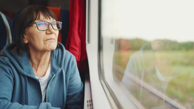 A pensive old woman in glasses rides on a train looking out the window and being reflected in it, Slow motion 