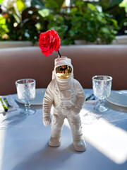 A unique astronaut figurine vase holding a vibrant red rose, set against a fresh, green leafy backdrop with elegant tableware