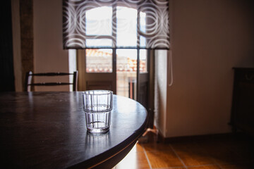 Empty glass cup on a wooden table and in the background an open door that leads to the outside of the house. Brooding environment.