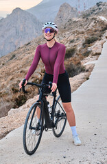 Fototapeta premium Happy professional female cyclist wearing cycling kit and helmet smiling at camera while standing with her bike outdoor with view on mountains. Sports motivation image. Good road for cycling. Spain.
