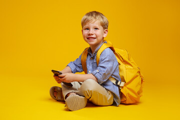 Joyful pupil boy in blue shirt with yellow backpack holding mobile phone in hands, smiling at...