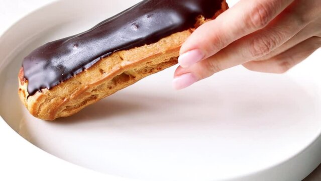 Chocolate eclair. Hand taking eclair from white plate. Close up of woman's hand with eclair. Woman's hand picking eclair with chocolate icing from white plate, glazed dessert close-up