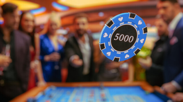 Elegantly Dressed Men and Women Enjoying Luxurious Atmosphere in a Casino Playing Roulette, Making Bets, Winning. Cinematic VFX Shot with Young Man Tossing a Casino Chip In Front of a Camera.