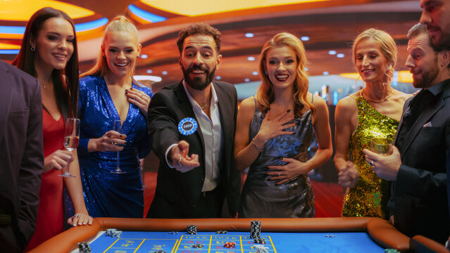 Elegantly Dressed Men and Women Enjoying Luxurious Atmosphere in a Casino Playing Roulette, Making Bets, Winning. Cinematic VFX Shot with a Young Man Tossing a Casino Chip In Front of a Camera.