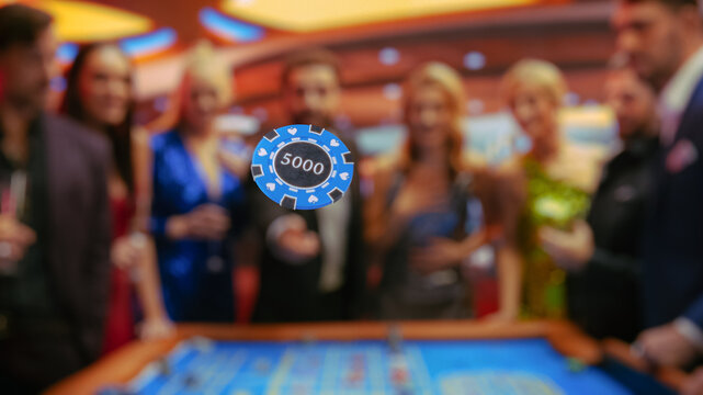 Cheerful Group of Young People Gathered Around a Roulette Table at a Modern Casino. A Handsome Hispanic Male Throwing a 5000 Dollar Casino Coin Towards the Camera, Crowd Cheering, and Having Fun.