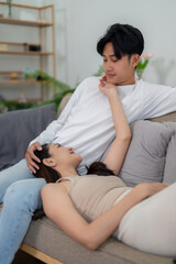 A young Asian couple sits comfortably on a beige couch, engaging in a pleasant conversation in a well-lit living room.
