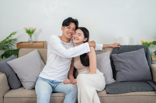 Cute Asian couple hugging and kissing couple's foreheads on the sofa. Sharing smiles and warm hugs in the comfort of their living room.