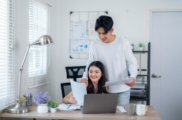 A smiling young Asian couple collaborates on a project, works together managing their budget, working on a laptop in a home office setting.