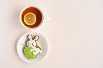 A gingerbread in the shape of a rabbit with an Easter egg on a plate and a cup of tea with a slice of lemon on a light table. Top view. Copy space