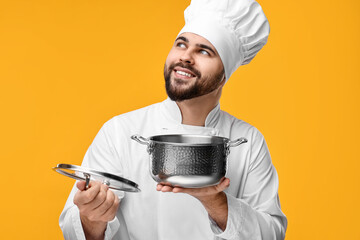 Happy young chef in uniform holding cooking pot on orange background