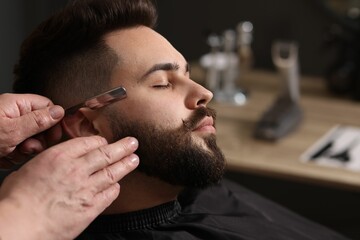 Professional barber shaving client's beard with blade in barbershop, closeup