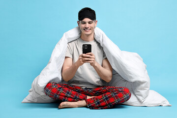Happy man in pyjama wrapped in blanket using smartphone on light blue background