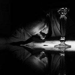 Vertical grayscale closeup of a man in the darkness looking at a glass goblet.