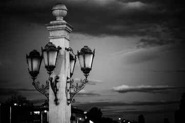 Grayscale shot of a column with vintage street lamps against the background of the cloudy sky.