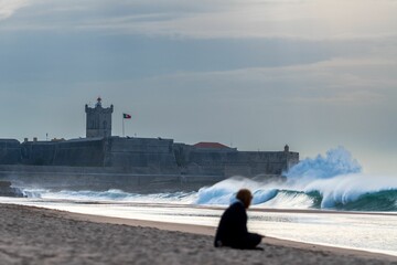 Caucasian female sitting on the seashore with strong waves in Portugal