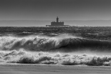 Grayscale shot of the strong ocean waves with a lighthouse in the background