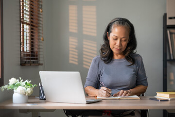 Serene and focused senior businesswoman writing notes in a notebook with a laptop open in her...