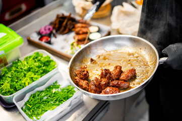 In a bustling professional kitchen, a chef expertly cooks meat in a sizzling pan. Fresh greens and prepared dishes surround the scene, creating an organized yet busy atmosphere