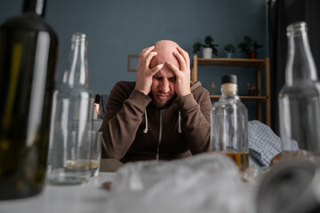 Alcohol addiction abuse and alcoholism concept. Upset millennial man drinker alcoholic sitting at home with empty bottles drinking whiskey alone, sad depressed addicted man having problem.
