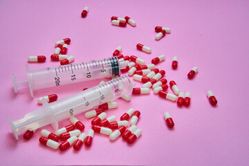 Two syringes and a pile of red and white pills and capsules on a pink table.