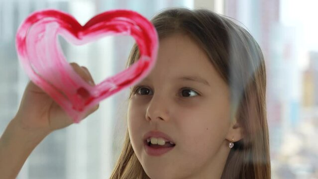 Cute girl painting heart on window with paintbrush, close-up