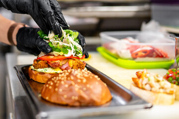 A chef in black gloves assembles a gourmet burger with fresh toppings on a bun, in a professional kitchen setting. The burger consists of lettuce, tomatoes, and other fresh ingredients.
