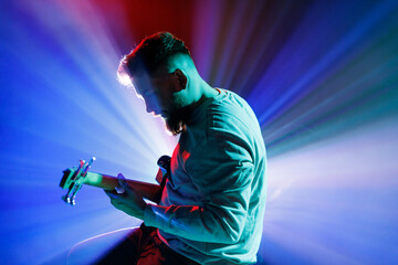 Side view portrait of young man performing playing electric guitar in neon light and visible smoke...