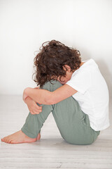 A young boy is seated on the wooden floor with head bowed down