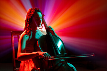 Portrait of female cellist performing classical melodies with radiant red and purple stage lights...