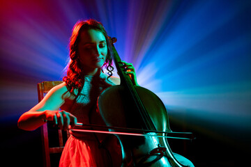 Portrait of female cellist performing classical melodies with radiant blue and purple stage lights behind her on stage. Concept of hobby and work, music festivals, concerts, symphony show, culture.