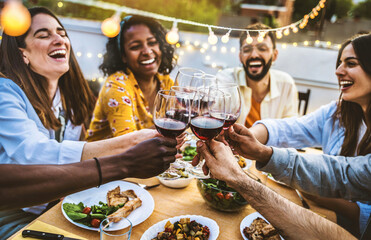 People toasting red wine glasses on rooftop dinner party - Happy friends eating meat and drinking wineglass at restaurant patio - Food and beverage lifestyle concept with guys and girls dining outdoor