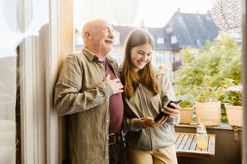 Happy senior man laughing while standing with teenage granddaughter holding smart phone in balcony