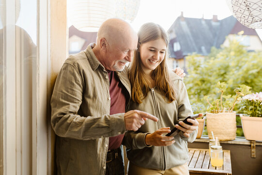 Smiling teenage girl sharing smart phone with grandfather standing in balcony