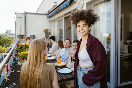 Portrait of smiling young woman with hand in pocket holding wineglass while celebrating dinner party with friends in bal