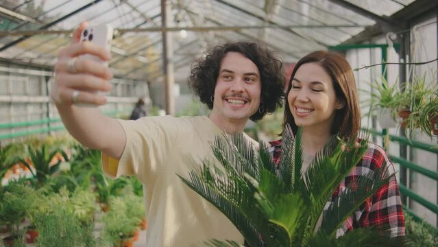 the couple chose a beautiful green plant and take a selfie with it on a smartphone and are happy. Funny couple. A man and a woman are photographed in a greenhouse