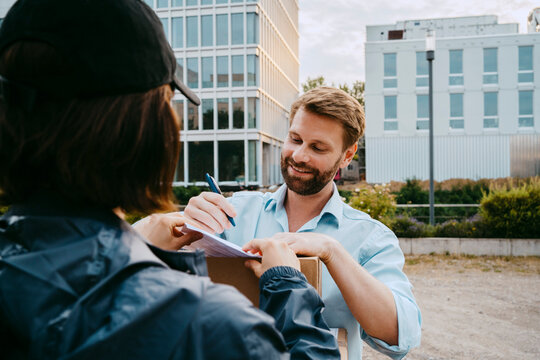 Smiling man signing on document while receiving parcel from delivery person