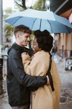 Side view of happy multiracial couple standing under umbrella at street