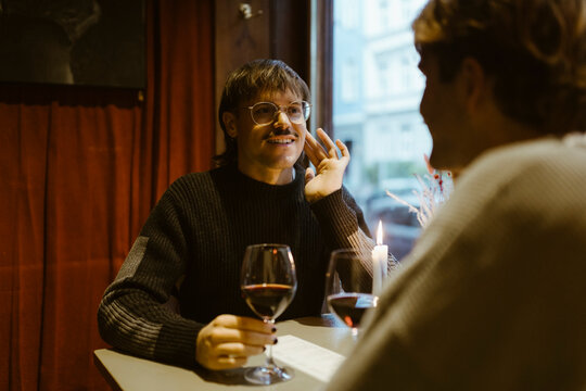Smiling gay man holding wineglass and talking with boyfriend while sitting at restaurant
