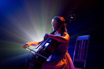 Talented young woman, cellist immersed in music, with striking halo of stage lights accentuating her silhouette on stage. Concept of hobby and work, music festivals, concerts, symphony show, culture. - obrazy, fototapety, plakaty