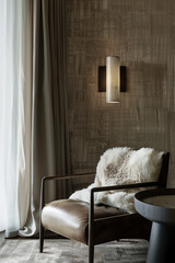 A modern wall lamp in the shape of an elongated cylinder with frosted glass and brass details, mounted on textured wallpaper in neutral tones. A plush sheepskin throw is draped over one armchair for w