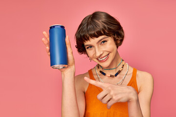 joyful young woman with short brunette hair pointing at soda can on pink, summer beverage