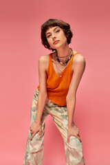 young woman in her 20s with short brunette hair posing in vibrant summer attire on pink backdrop