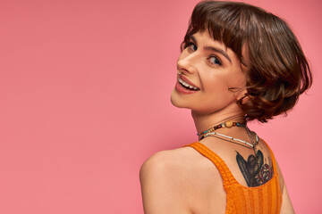 happy tattooed woman in 20s with nose piercing and brunette short hair smiling on pink background