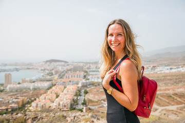 A woman is smiling and holding a red backpack while standing on a hill