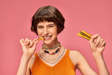 young pierced woman in her 20s biting sweet and sour gummy stripe and holding candies on pink