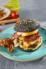 Gourmet black burger with bacon and crispy onions on a turquoise plate, gray textured backdrop with a hint of sunlight