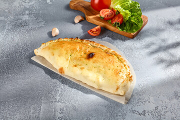 Golden calzone pizza on parchment paper, with fresh tomatoes and garlic in the background on a textured grey surface - 776110946
