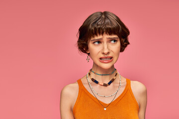 puzzled young girl in her 20s standing in orange knitted tank top on pink background, unsure