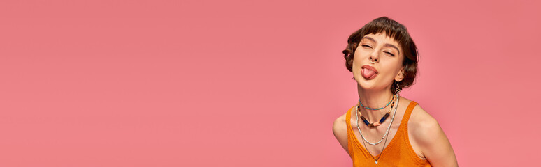 cheeky playful girl in 20s with short brunette hair sticking tongue out on pink background, banner