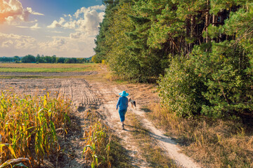 A young woman with a dog walks along a dirt road between a field and a forest. Drone view